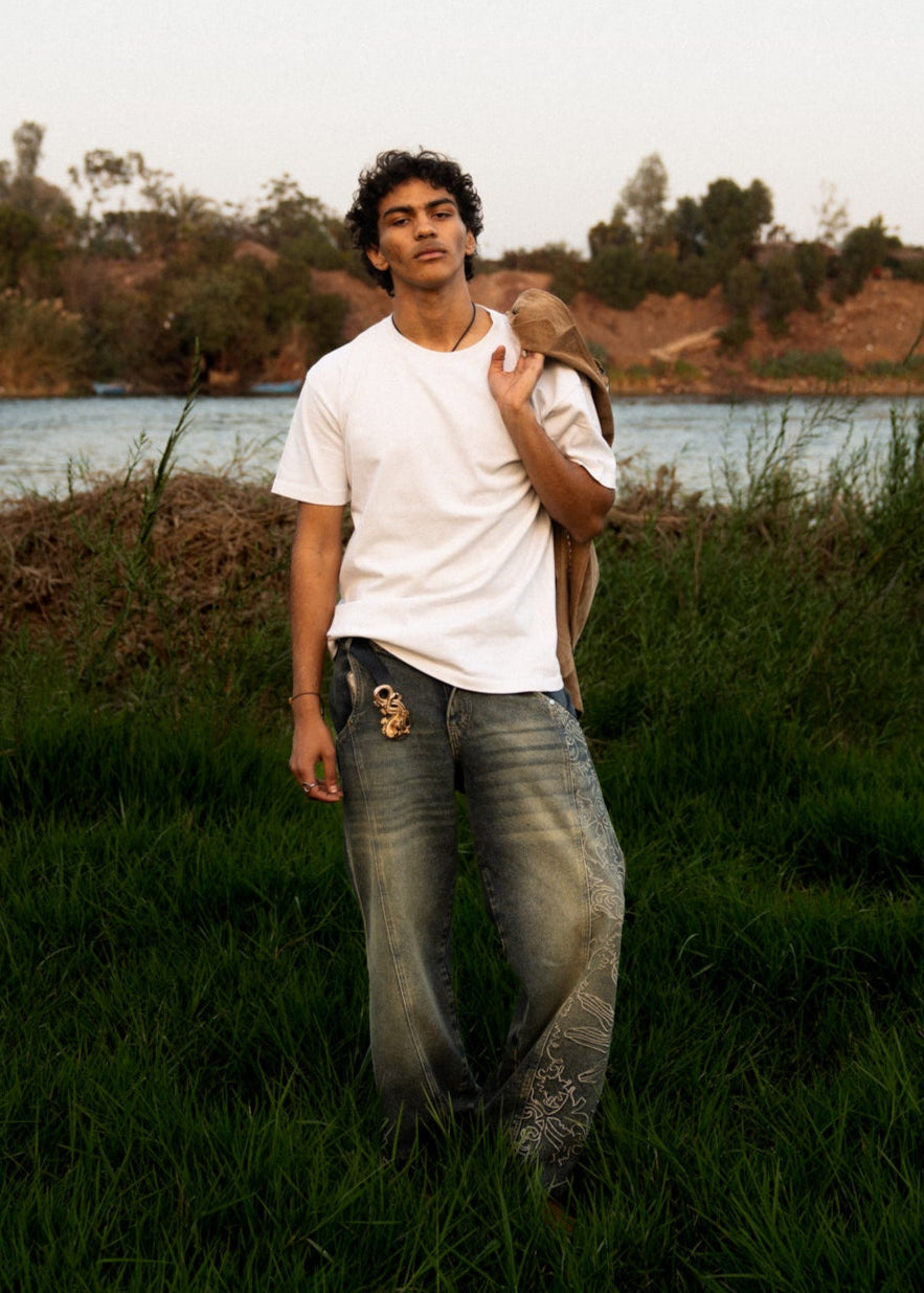 A person standing in a natural setting wearing a white t-shirt and blue denim jeans with embroidery on the back pocket.