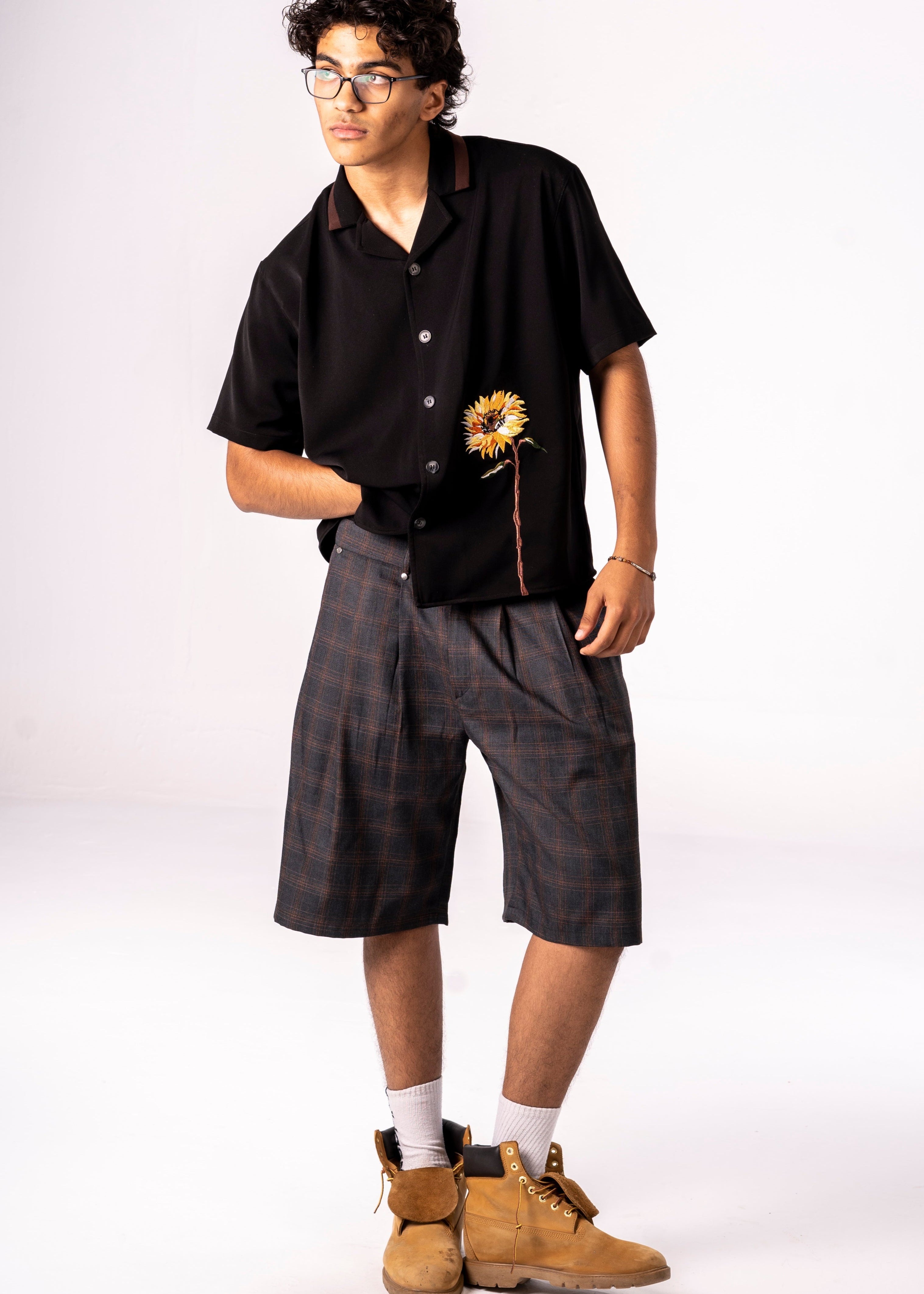 Person wearing a black shirt with a sunflower design, gray shorts, and brown boots on a white background