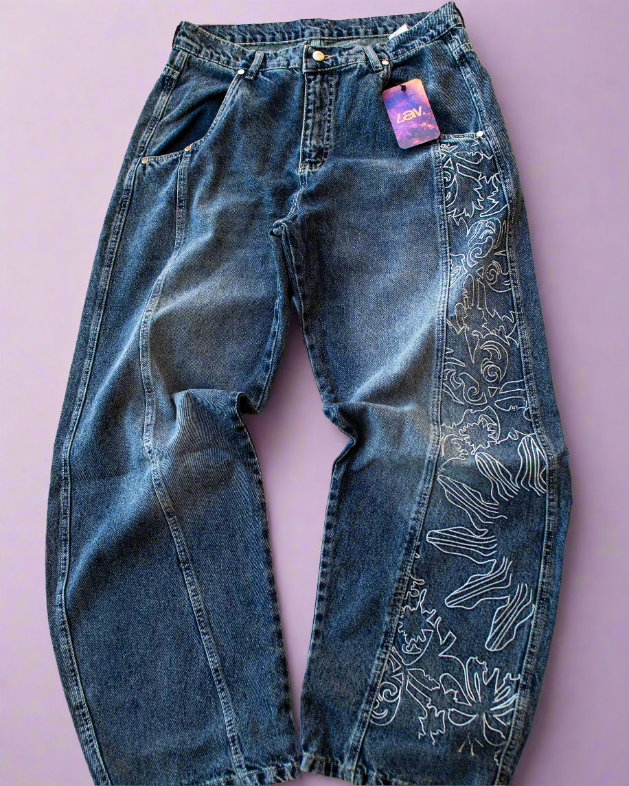 A pair of blue denim jeans with full-length embroidery on one leg, displayed against a purple background.